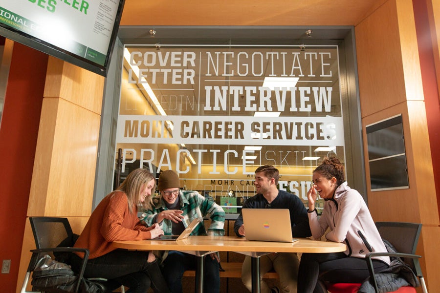 A group of students sitting, talking, and laughing around a table. Behind them is a glass wall with various sayings on it, including Career Center