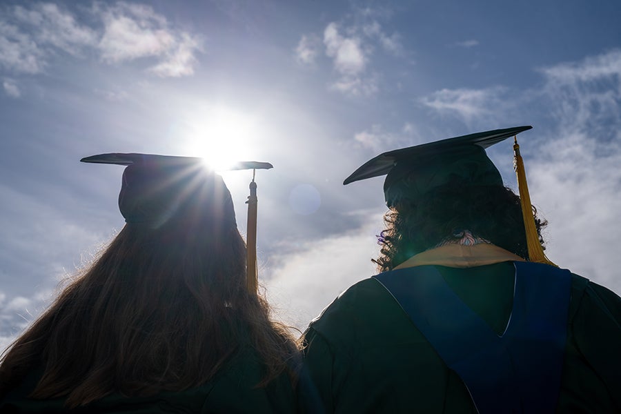 Two UO graduates in cap and gown with their backs turned to the camera, with a sunrise in front of them