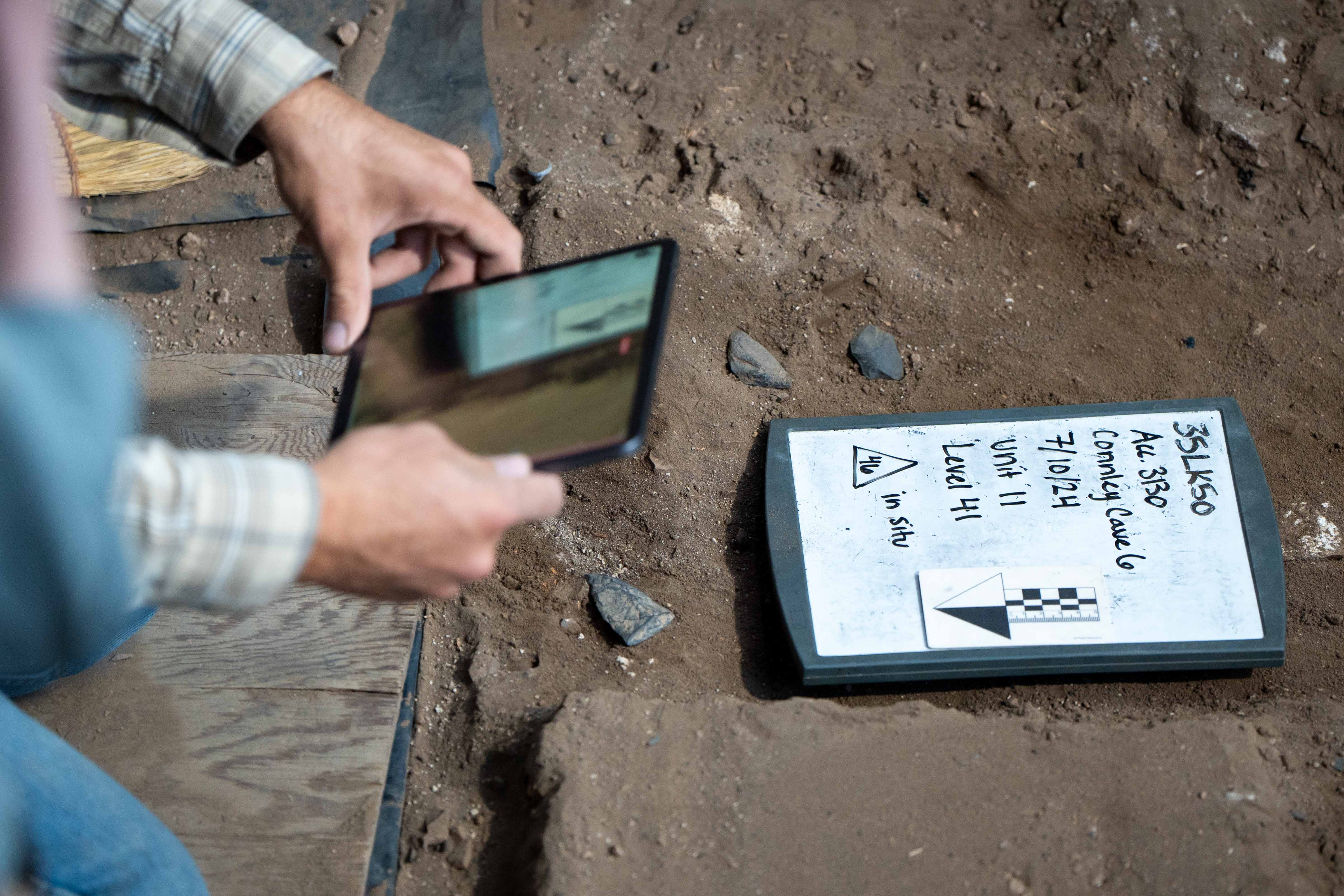 A person holding an ipad and taking a photo of the site and a board with notes