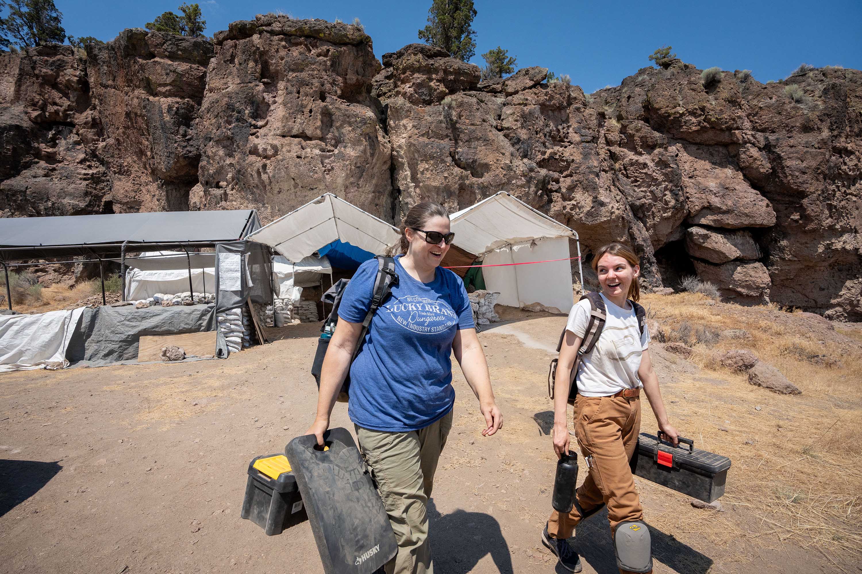 Stephanie Alexander and Gabi Gauthier walking outside in front of the site tents wearing backpacks and carrying toolboxes