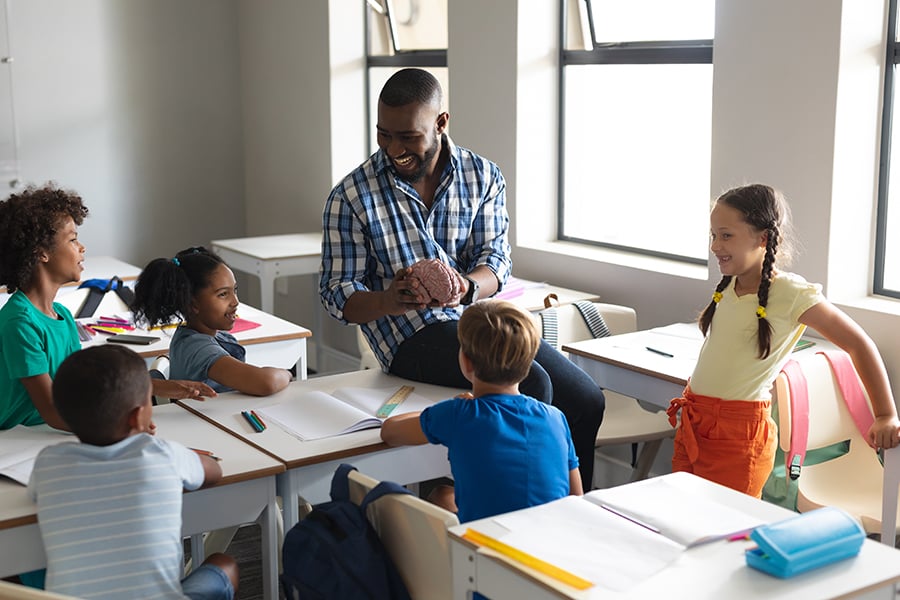 In a classroom, a man sits on the edge of a student desk and holds a brain. Young elementary students are at their desks looking at him.