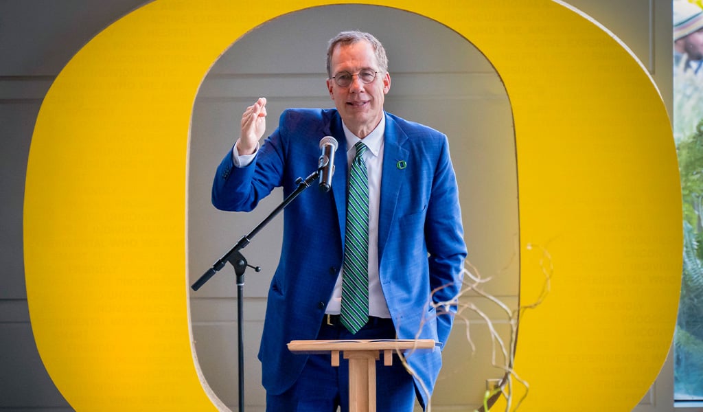 Oregon President Karl Scholz addressing a crowd at the Ford Alumni Center, speaking at a podium in front of a large Oregon ‘O’ statue.