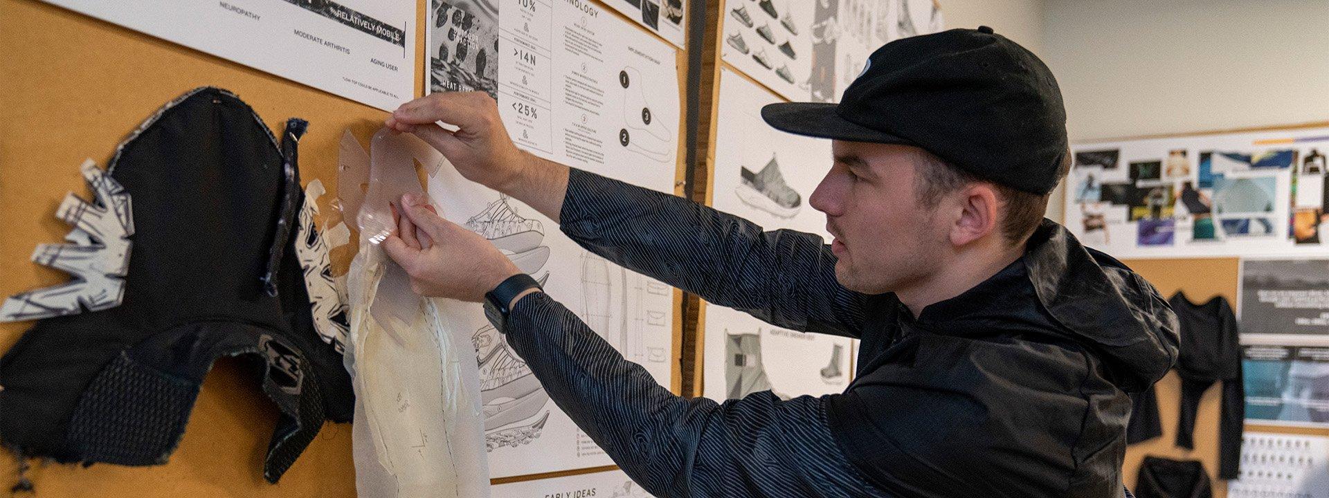 A UO student wearing a black baseball cap pins materials onto a board with sneaker designs displayed across it.
