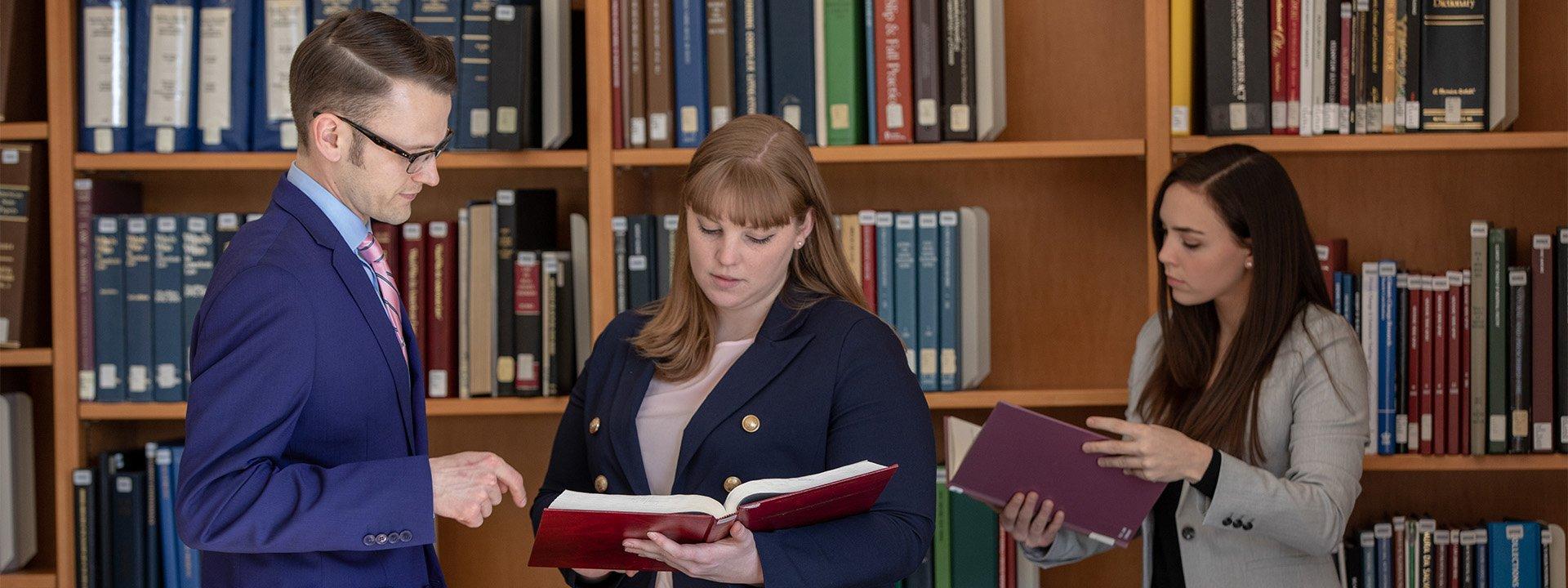 Formally dressed UO students looking at open books, with a shelf full of books in the background