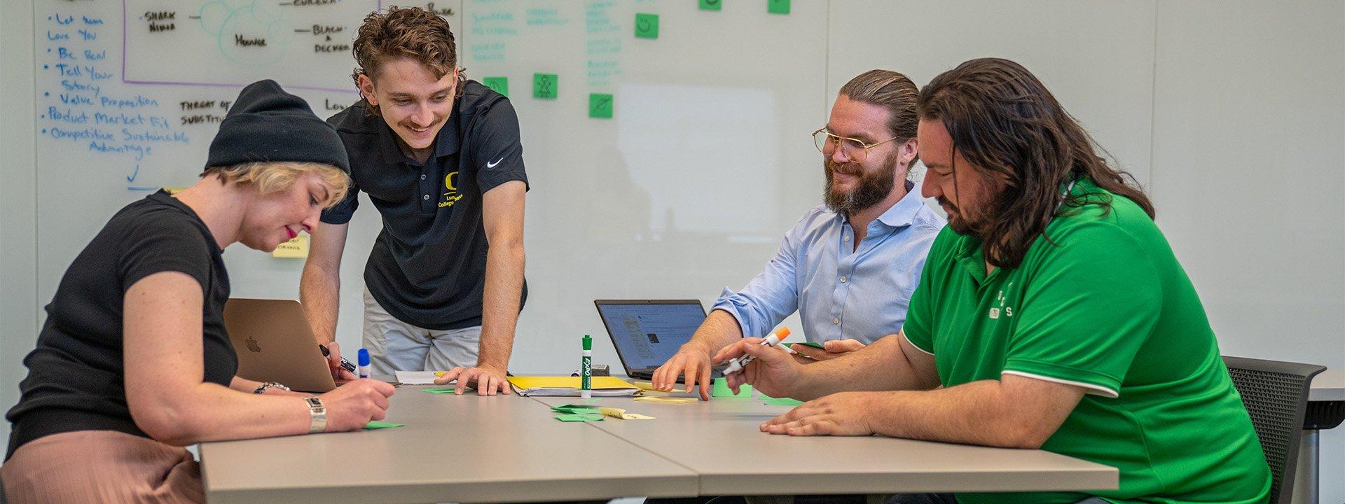 Four UO students sit at a table, working together, with a white board with writing on it in the background