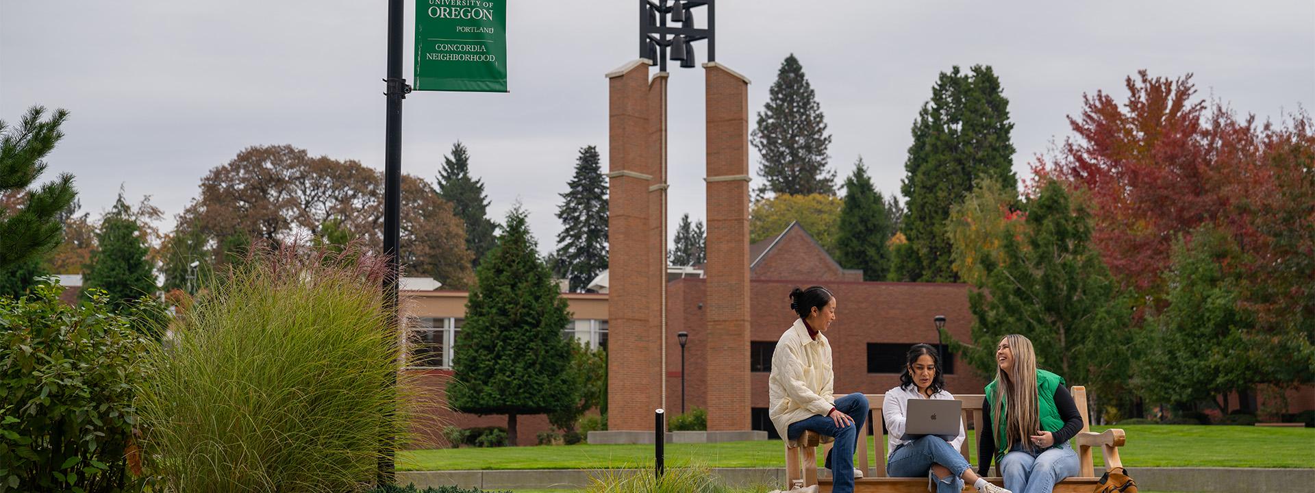 Three UO students sit on a bench on campus. One looks at a laptop while the other two laugh.