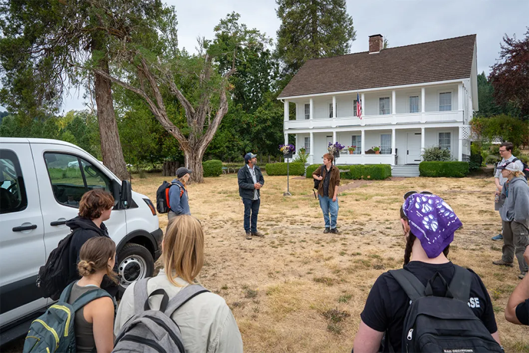 Exterior shot of Applegate House, a 2-story white house, and students standing in a circle outside it