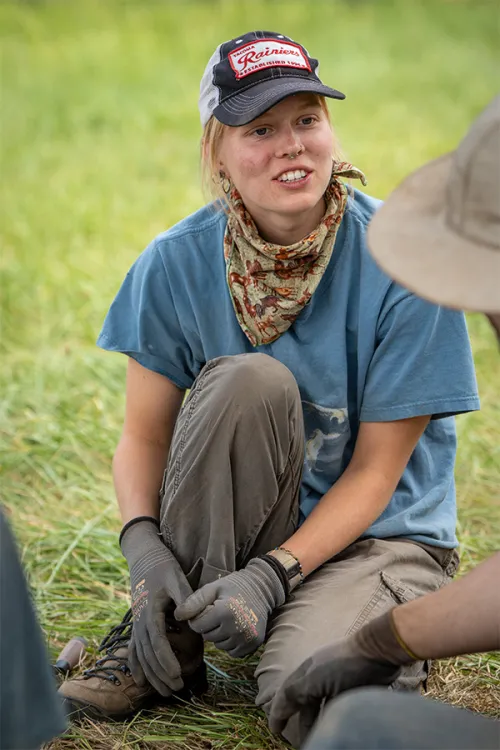 Addie, blonde white student, crouching on the grass, in work gloves and a bandana