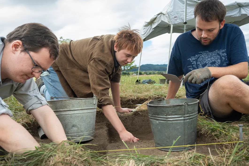 3 students with metal buckets digging in the dirt outside