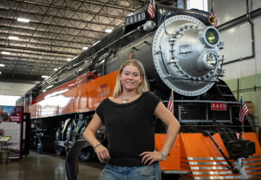 Chloe Mills stands in the foreground, with a historic train engine car behind her. 