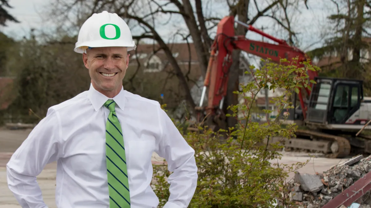 A man in a white shirt and green tie is wearing a construction hard hat with the UO logo on it. He smiles at the camera, with a claw claw excavator in the background