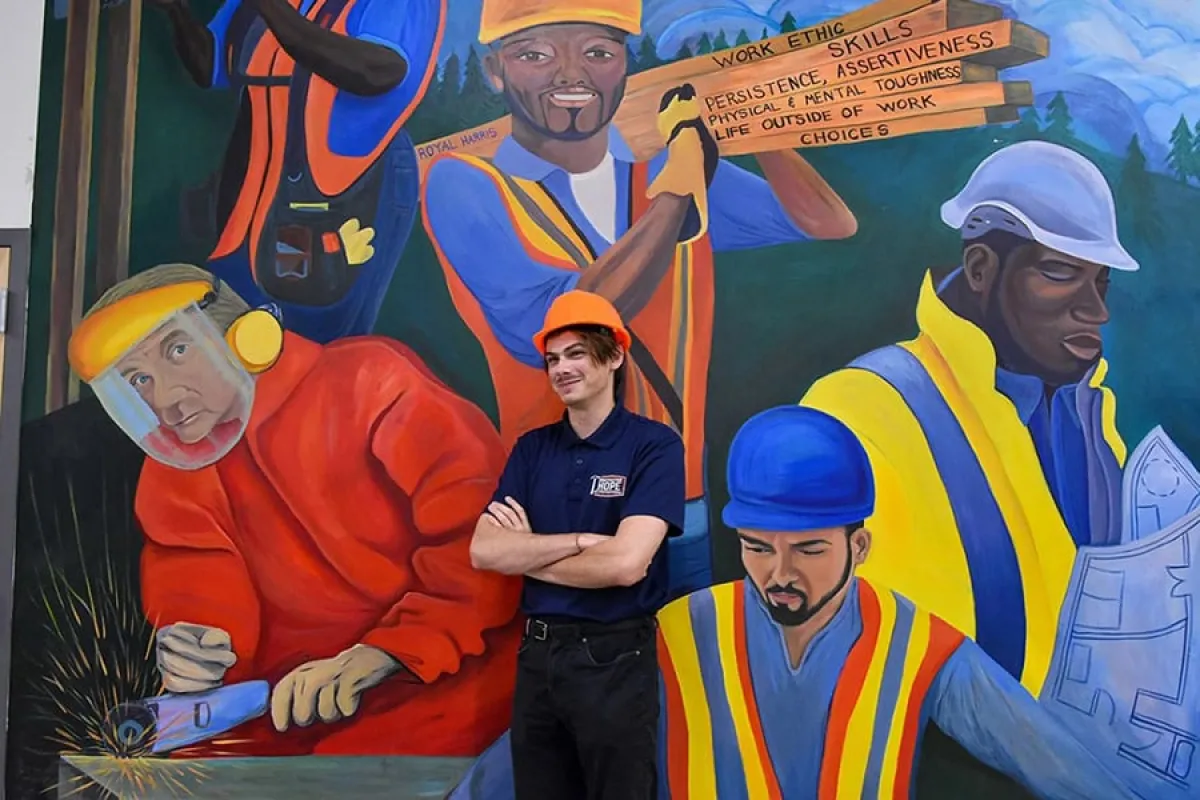 A UO student in a hard hat stands in front of a mural depicting construction workers