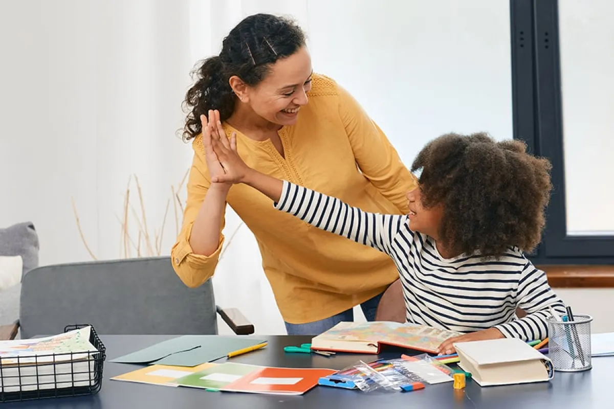 A smiling woman high-five's a child seated at a table, with notebooks and pencils spread out on the table
