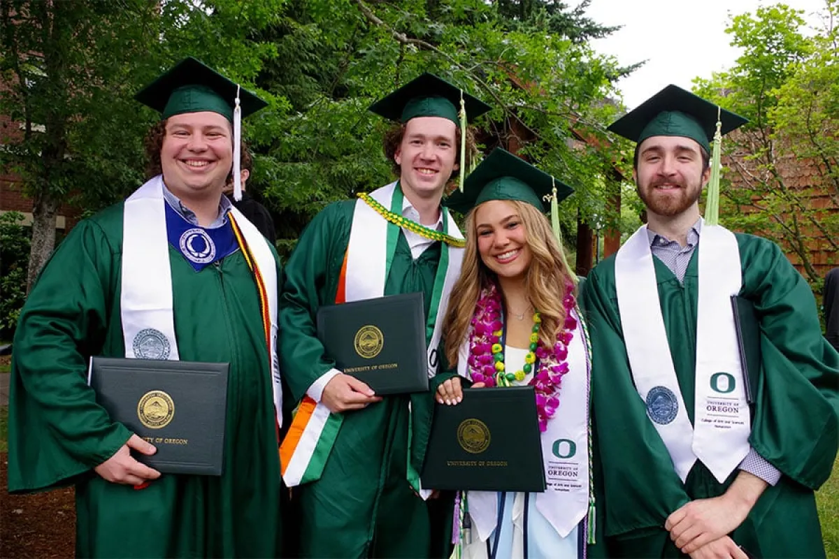 Four UO graduate students in green caps and gowns pose for a photo with their degrees