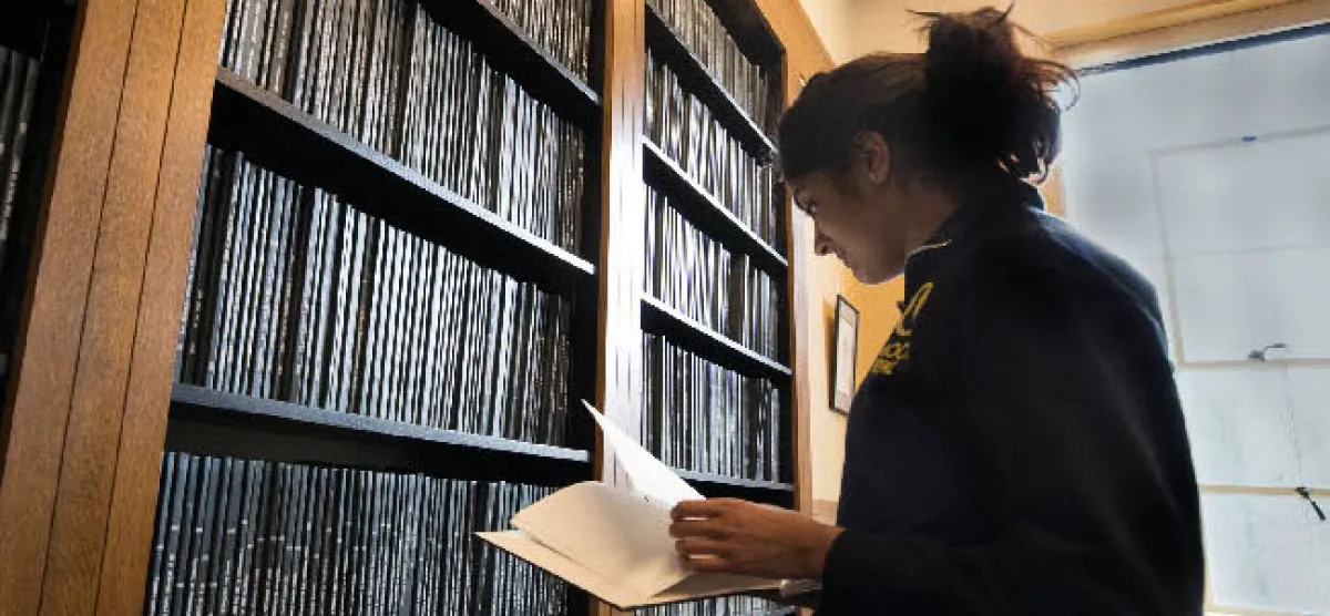 A student flips through a book in front of a filled bookcase 