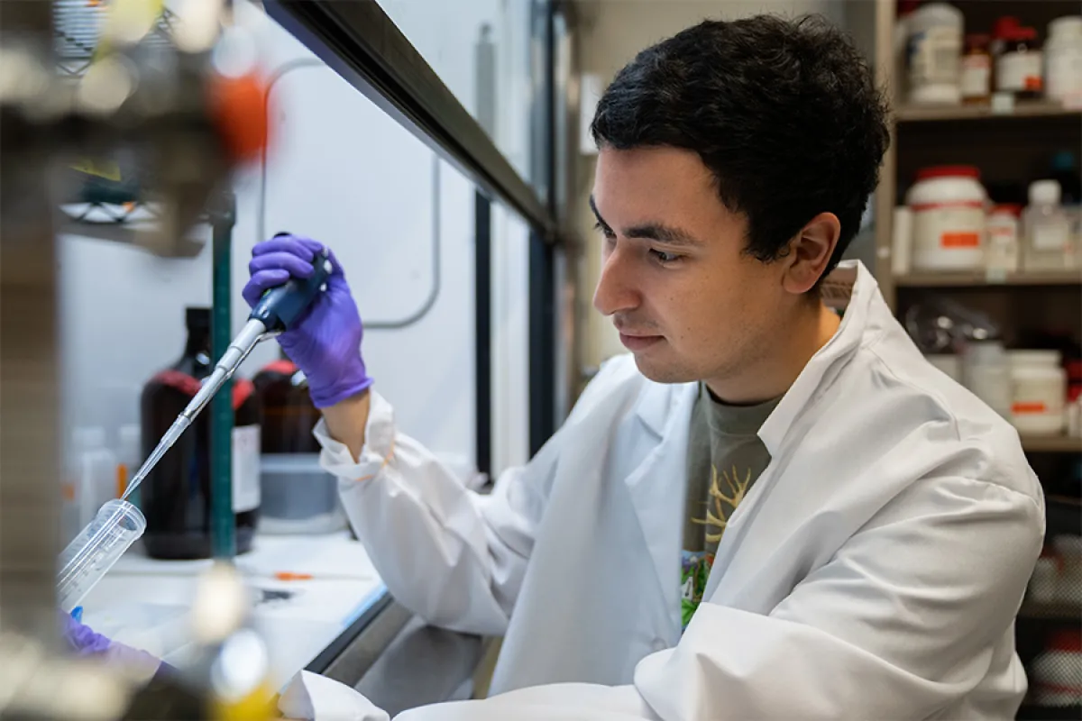 A UO student wearing a white lab coat and rubber gloves uses a pipette in a chemistry lab