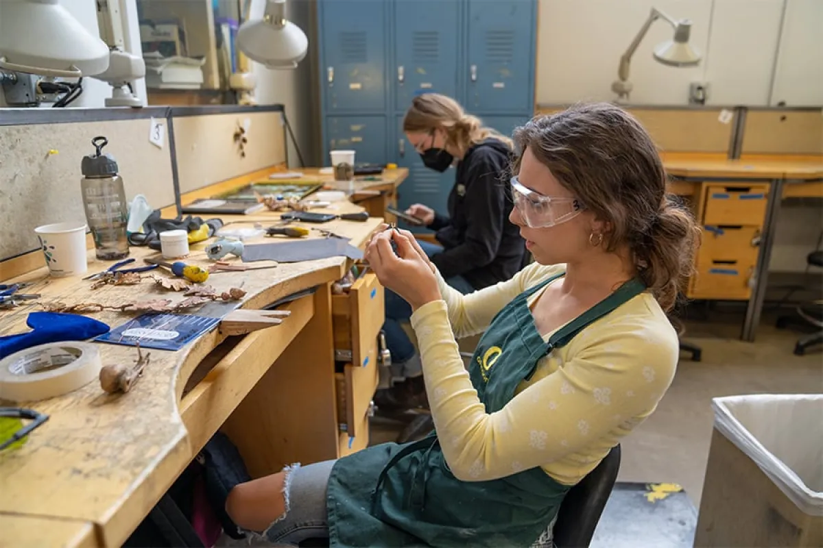 Two UO students work in a design workshop, sitting at wooden tables covered with tools and materials