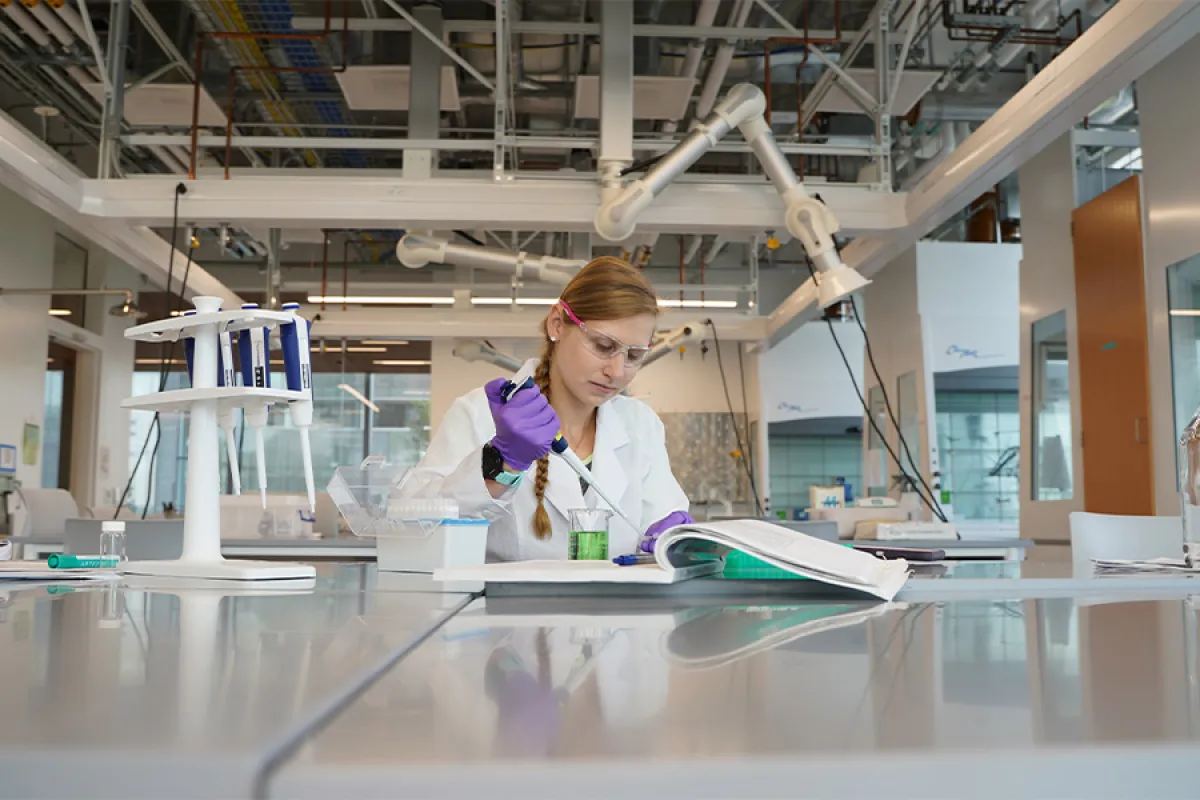 A student, wearing a white lab coat, rubber gloves and safety glasses, sits in a lab, using a pipette to transfer materials