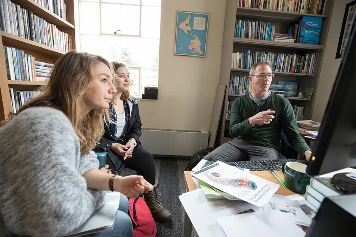 Two students and a UO faculty member sit in an office, looking off at a computer screen out of frame.