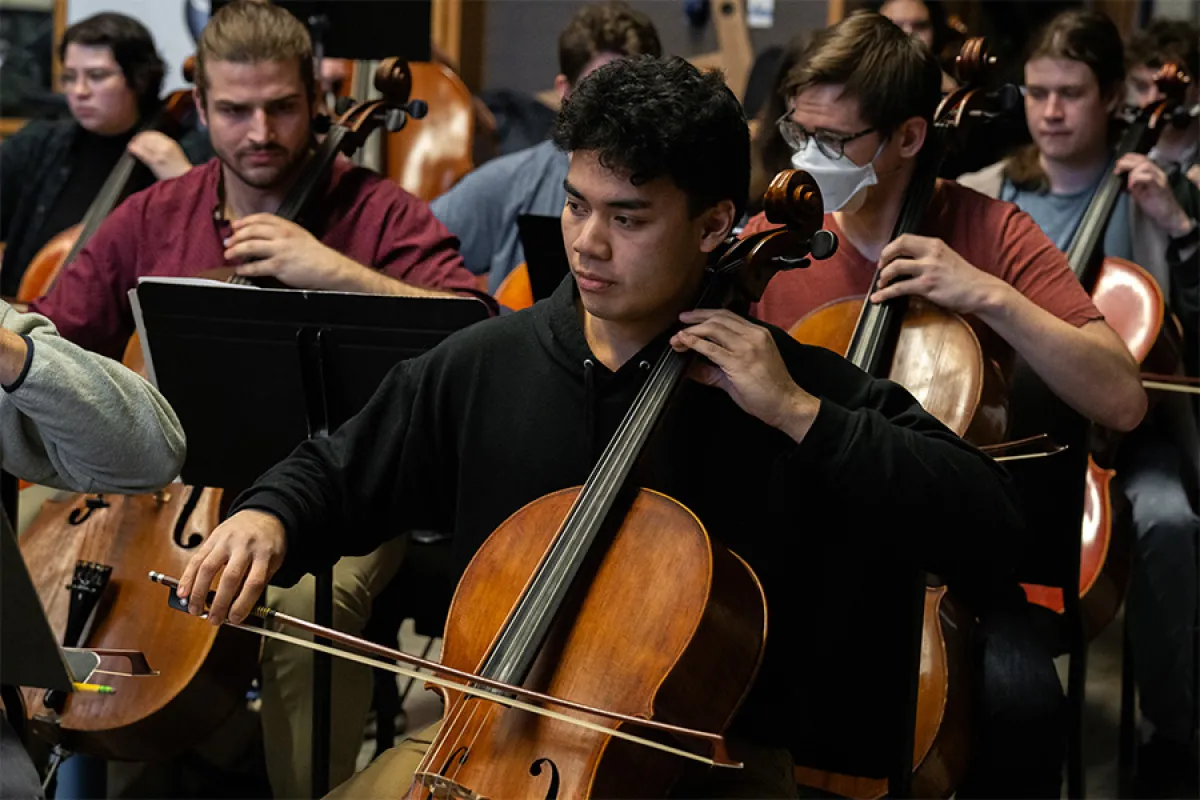 A UO student, playing a cello, sits in orchestra rehearsal, surrounded by other students