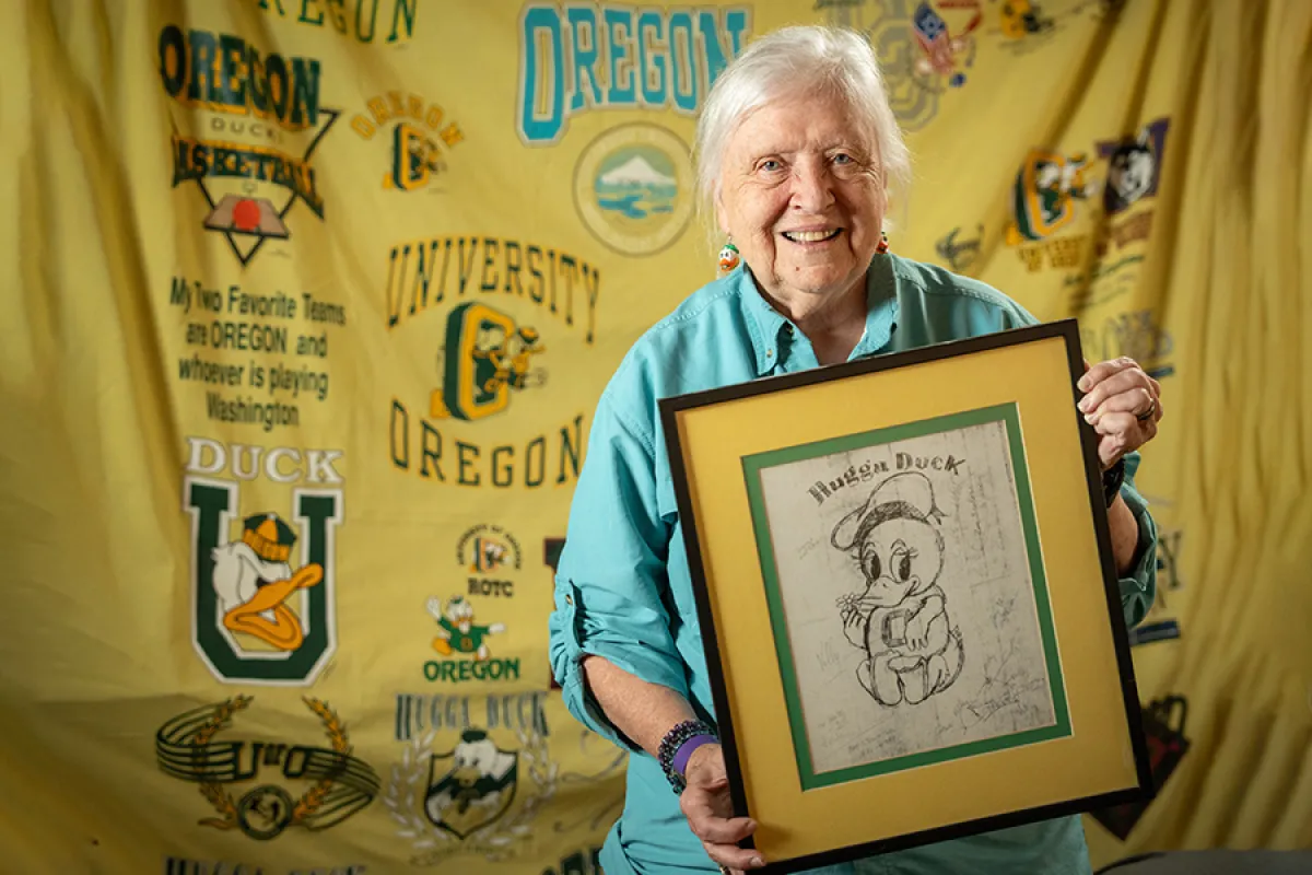 Elderly white woman in front of a yellow backdrop with a myriad of UO logos. She is holding a framed drawing of the Hugga Duck.
