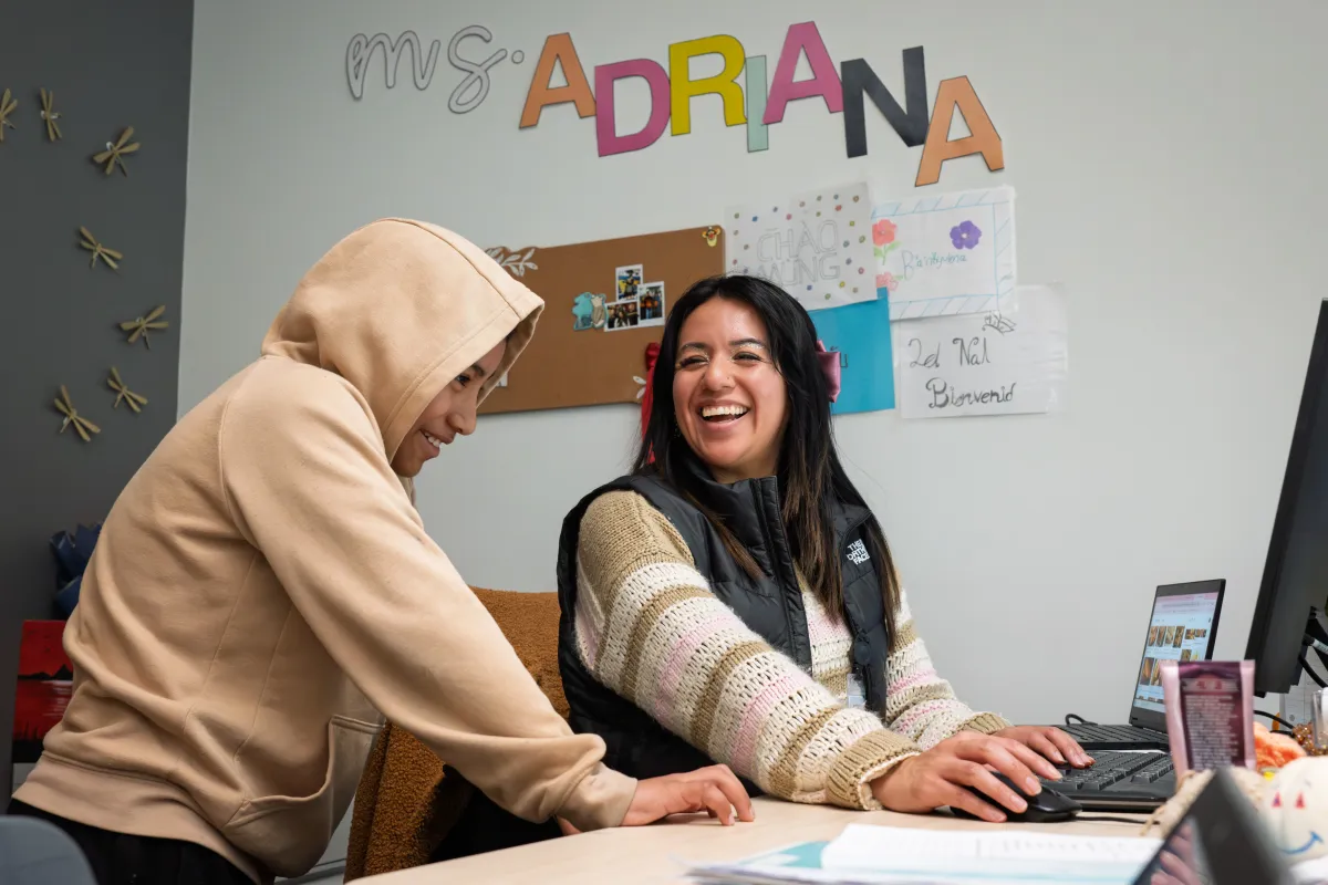 Adriana Alvarez smiles as she speaks with a student wearing a hoodie in a classroom. 