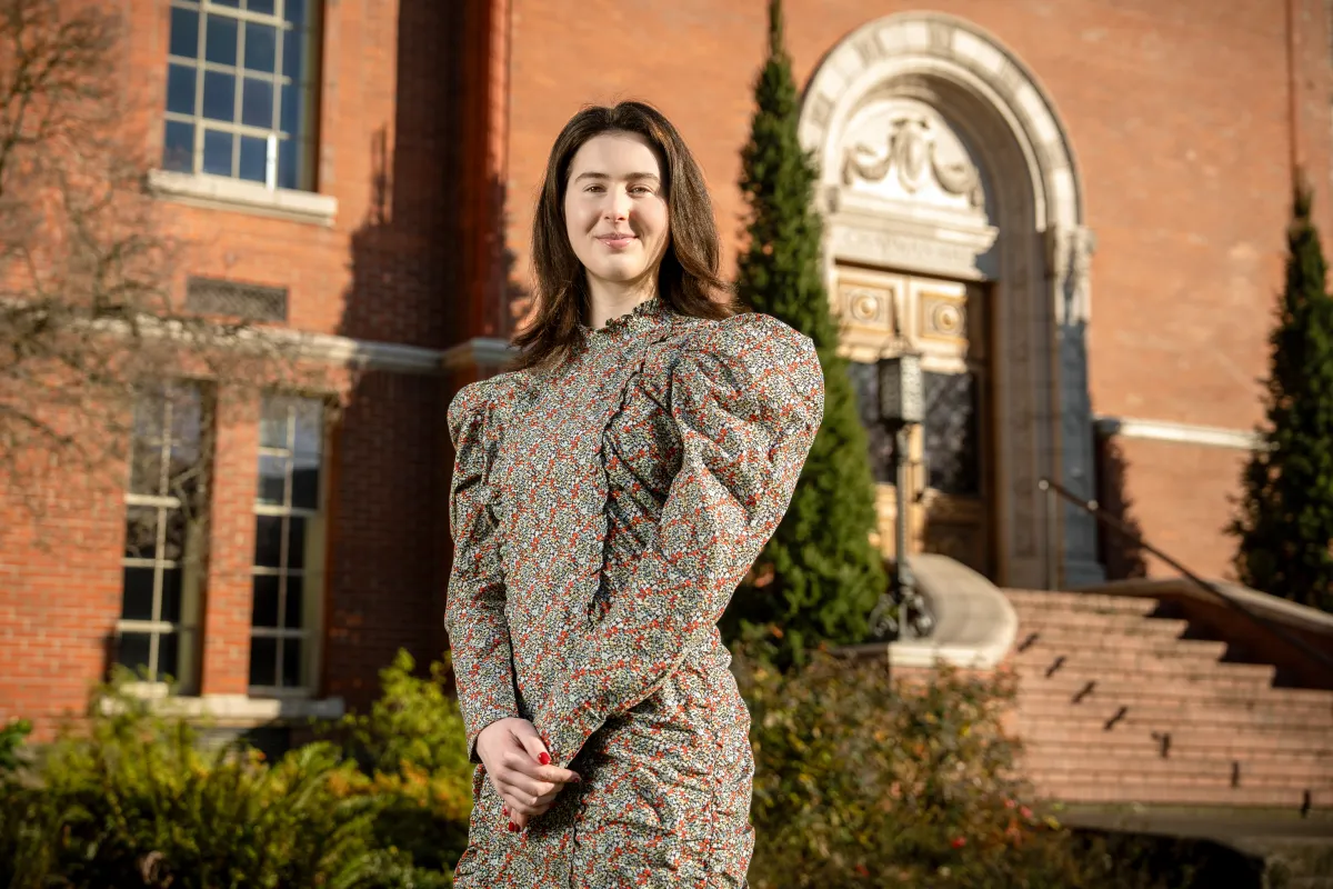 Beatrice standing infront of a brick building, looking at the camera and smiling 